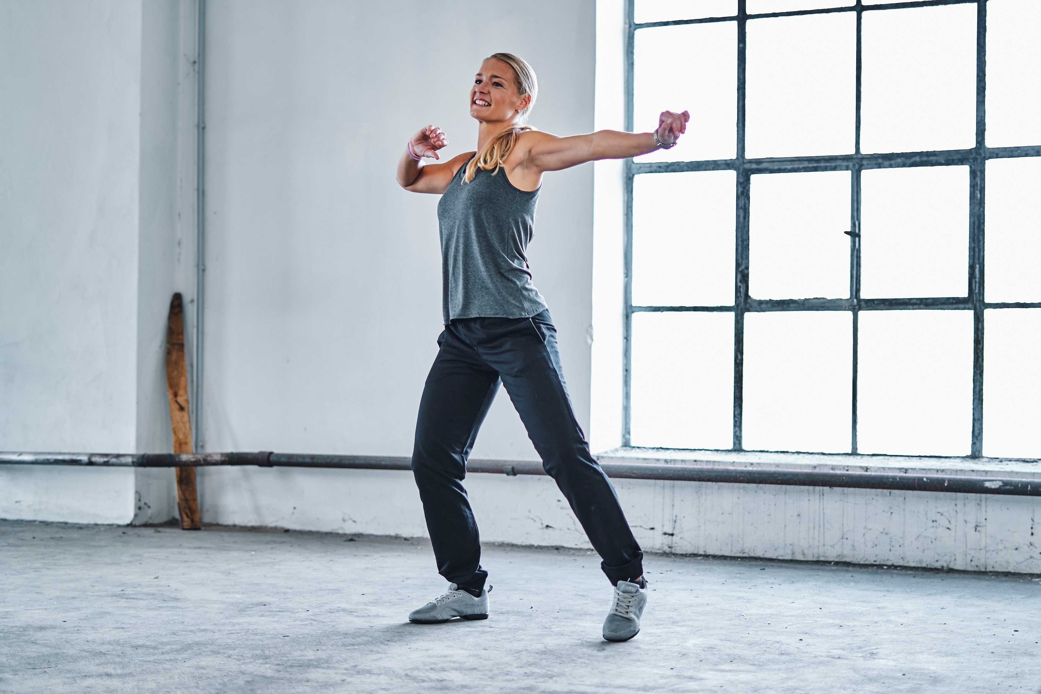 Person in Sportkleidung streckt die Arme in einem geräumigen Raum im Industriestil mit einem großen Fenster.